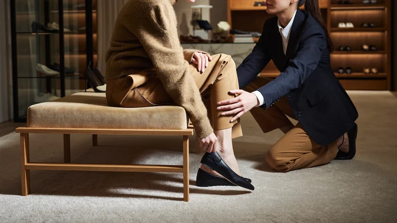 Ilustrace zákazníka zkoušejícího botu na nízké lavici za asistence prodavače
